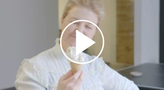 A person smiling with eyes closed, holding an ice cream cone with a large scoop. They are wearing a light-colored, ruffled blouse. The background includes a blurred interior setting.