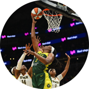 A basketball player in a green and yellow uniform jumps to make a layup as two defenders in white "Fever" jerseys attempt to block her shot near the hoop during a game.