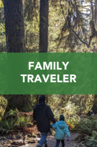 An adult and a child, bundled in winter jackets, walk hand in hand on a forest trail with tall trees. A green banner across the image reads "Family Traveler" in bold white letters.