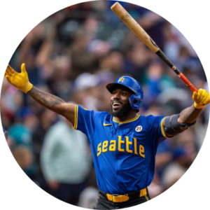 A baseball player in a blue "Seattle" jersey celebrates with arms wide open, holding a bat in one hand. The crowd is blurred in the background.