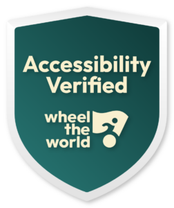 Shield-shaped badge with a dark green background displaying the text "Accessibility Verified" above "wheel the world" in a stylized font with a graphic of a person in a wheelchair.