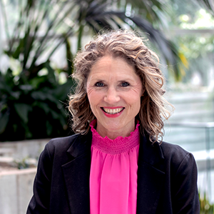 A person with curly hair smiles while wearing a black blazer over a bright pink top. They stand in front of a blurred background featuring indoor greenery.
