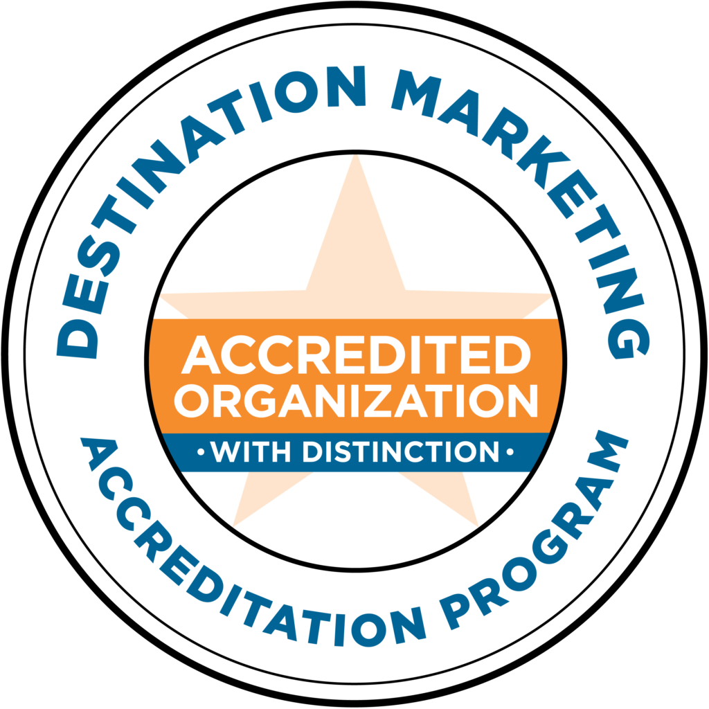 A circular badge with a star in the center reads “Accredited Organization With Distinction.” The outer ring says “Destination Marketing Accreditation Program.” Colors used are blue, orange, and white.