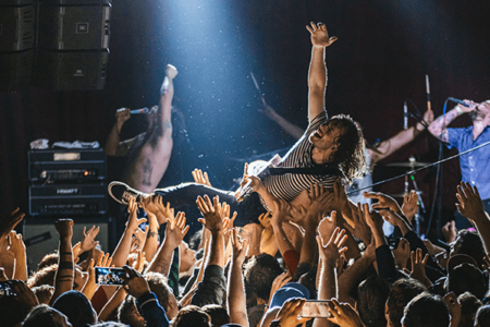 A rock concert scene with a crowd surfing performer in a striped shirt being held up by the audience. People around are cheering, with raised hands and some taking photos. The stage is dimly lit with a bright spotlight on the performer.