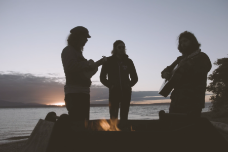 Three people silhouetted against a sunset sky play instruments around a campfire on a beach. One strums a guitar, another plays a ukulele, and the third stands between them. The scene is peaceful with an ocean in the background.