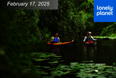 Two people kayaking on a calm, tree-lined river with green foliage and lily pads.
