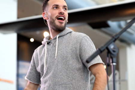 A man wearing a gray hoodie is performing on stage, holding a microphone. His expression is enthusiastic, with a slightly open mouth. There are blurred lights and industrial elements in the background.