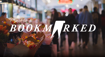 An indoor market scene with bouquets of colorful flowers on the left, blurred shoppers in the background, and the text "Seattle BOOKMARKED" prominently displayed over the image.