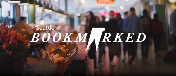 An indoor market scene with bouquets of colorful flowers on the left, blurred shoppers in the background, and the text "Seattle BOOKMARKED" prominently displayed over the image.