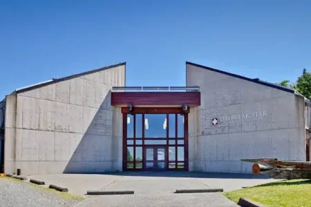 A modern building with angular concrete walls and large glass entrance under a clear blue sky. A wooden canoe is displayed on the right side, and lush green trees are visible in the background.